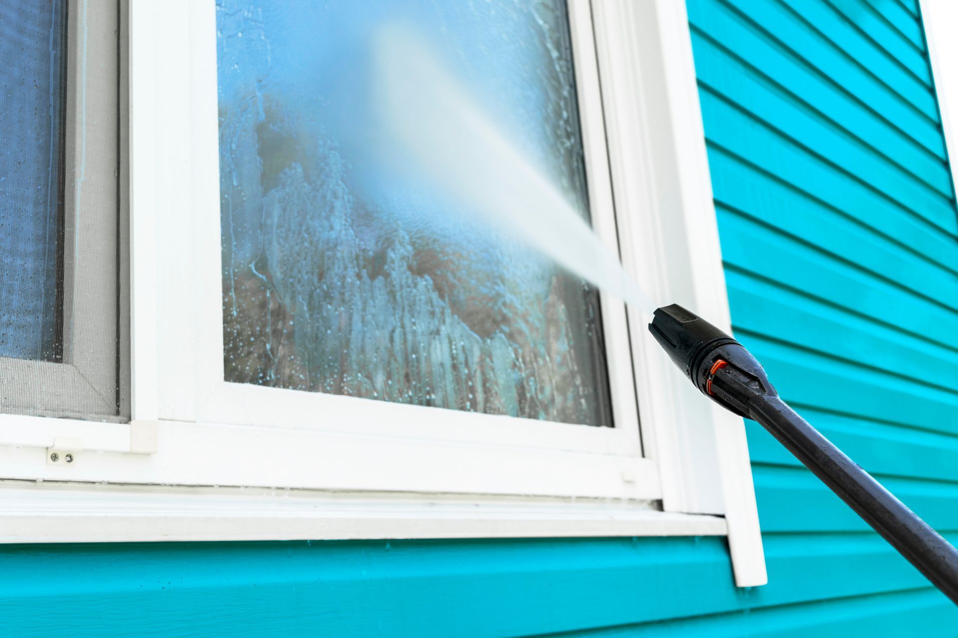 A person uses a pressure washer to clean a dirty window on a teal house