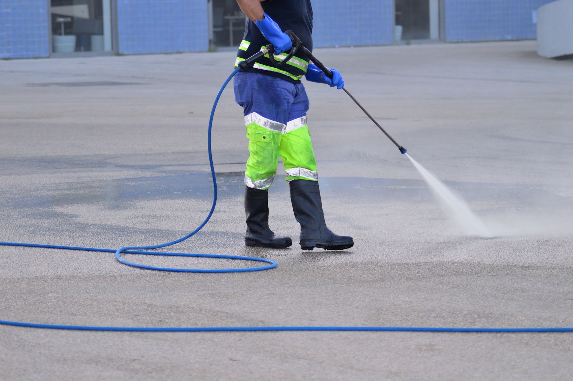 Person power washing a concrete surface with blue hose, wearing protective gear