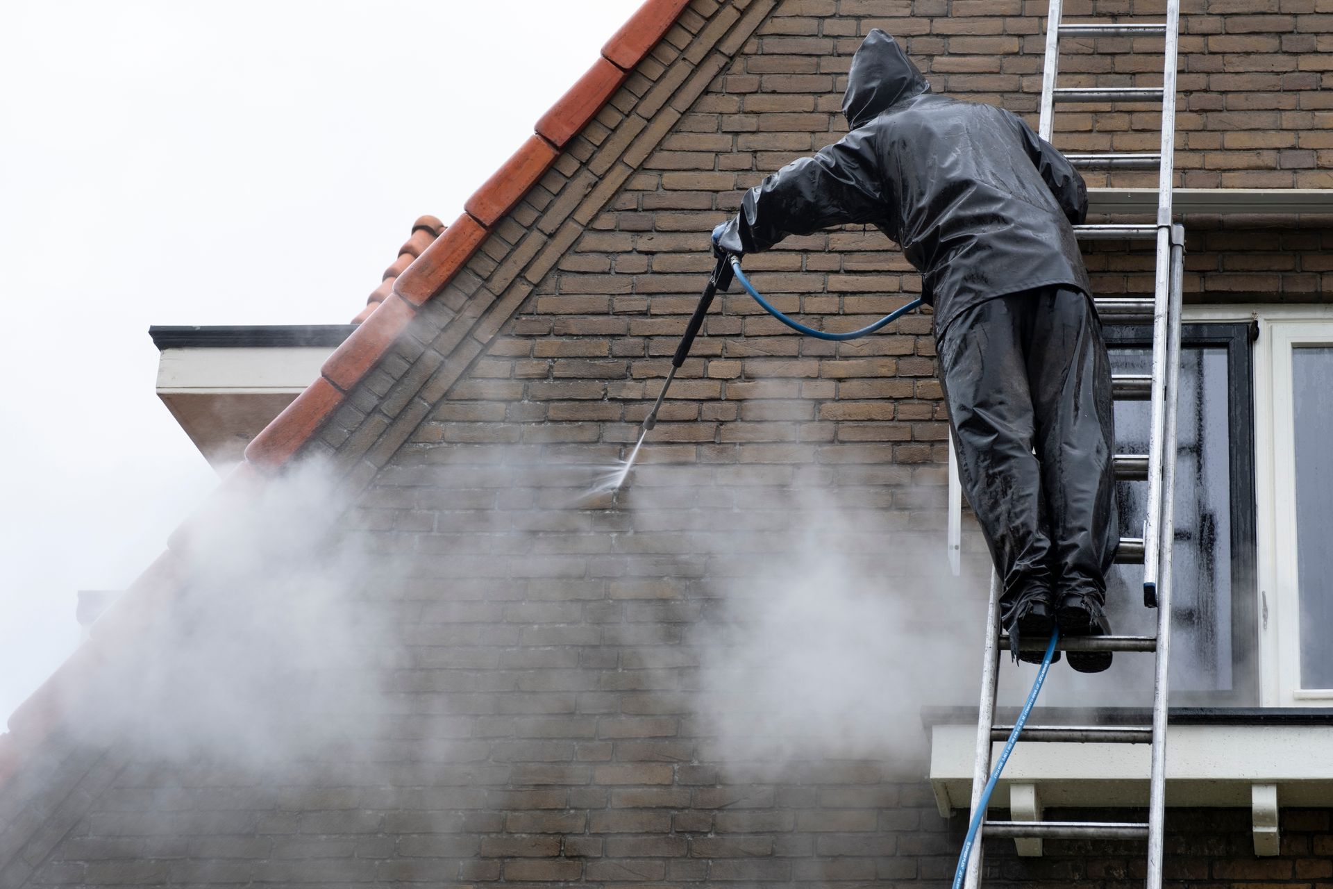 Person in rain gear power washing a brick house from a ladder