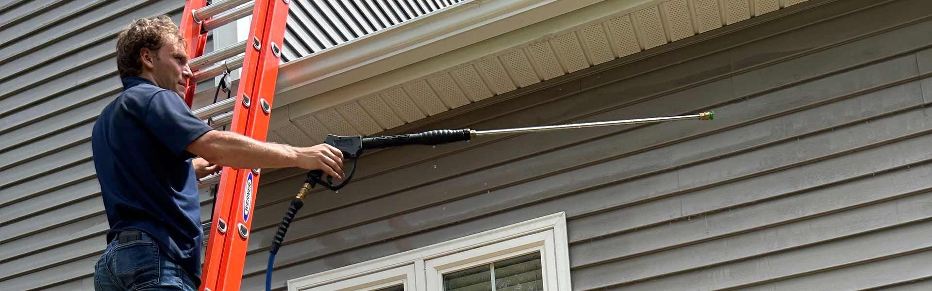 A person on a ladder using a power washer to clean the siding of a house