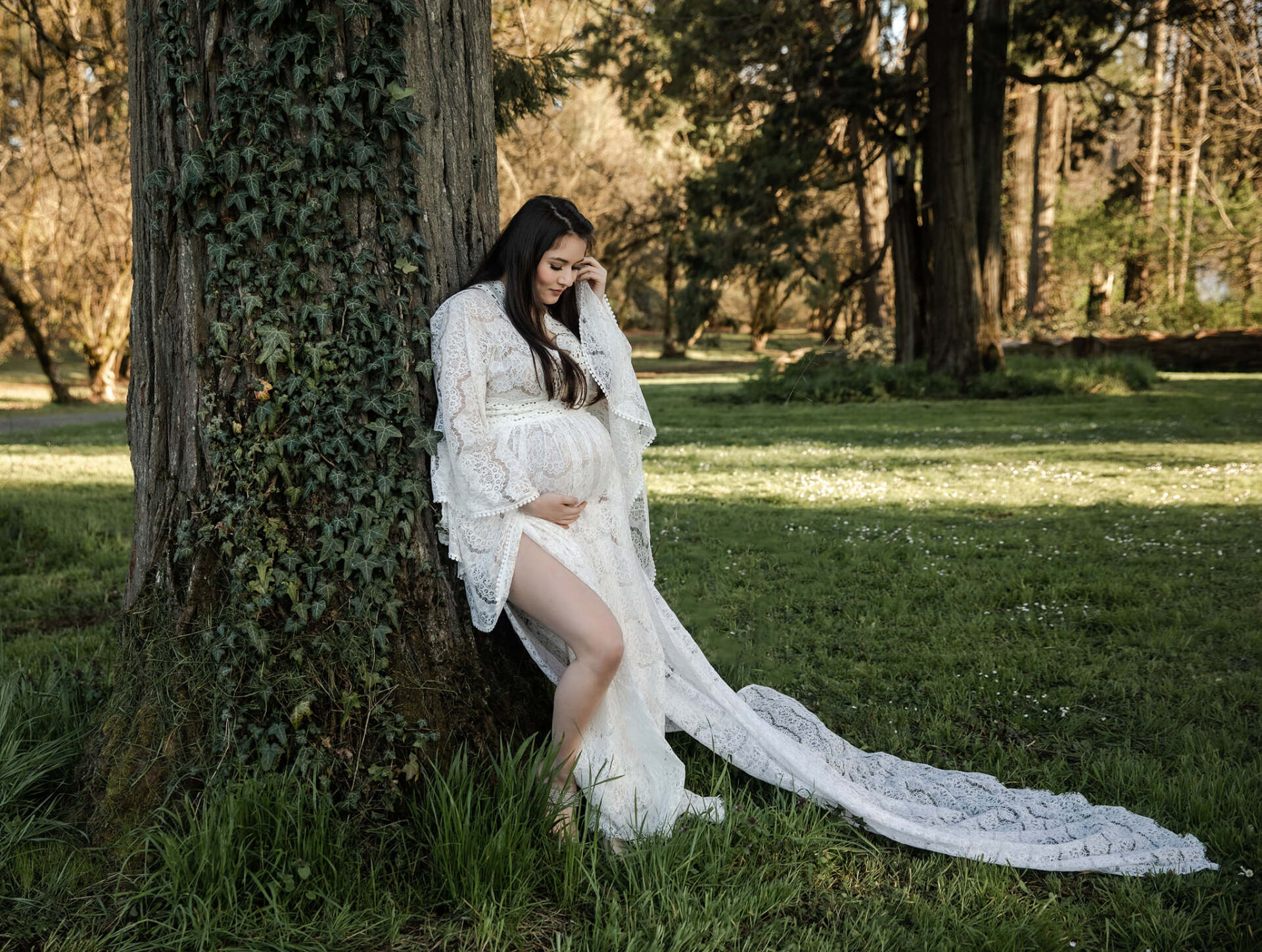 A pregnant woman in a white dress is leaning against a tree in a park.