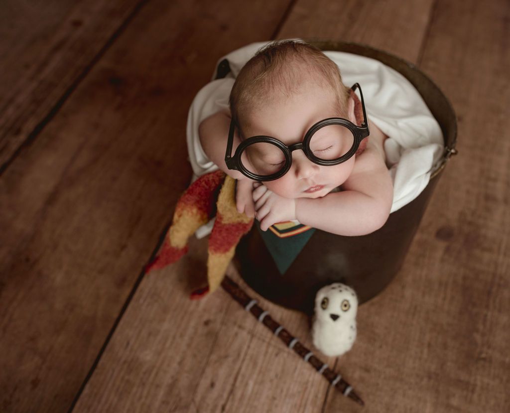 A newborn baby wearing glasses and a scarf is sleeping in a bucket. Themed Baby Photo sessions