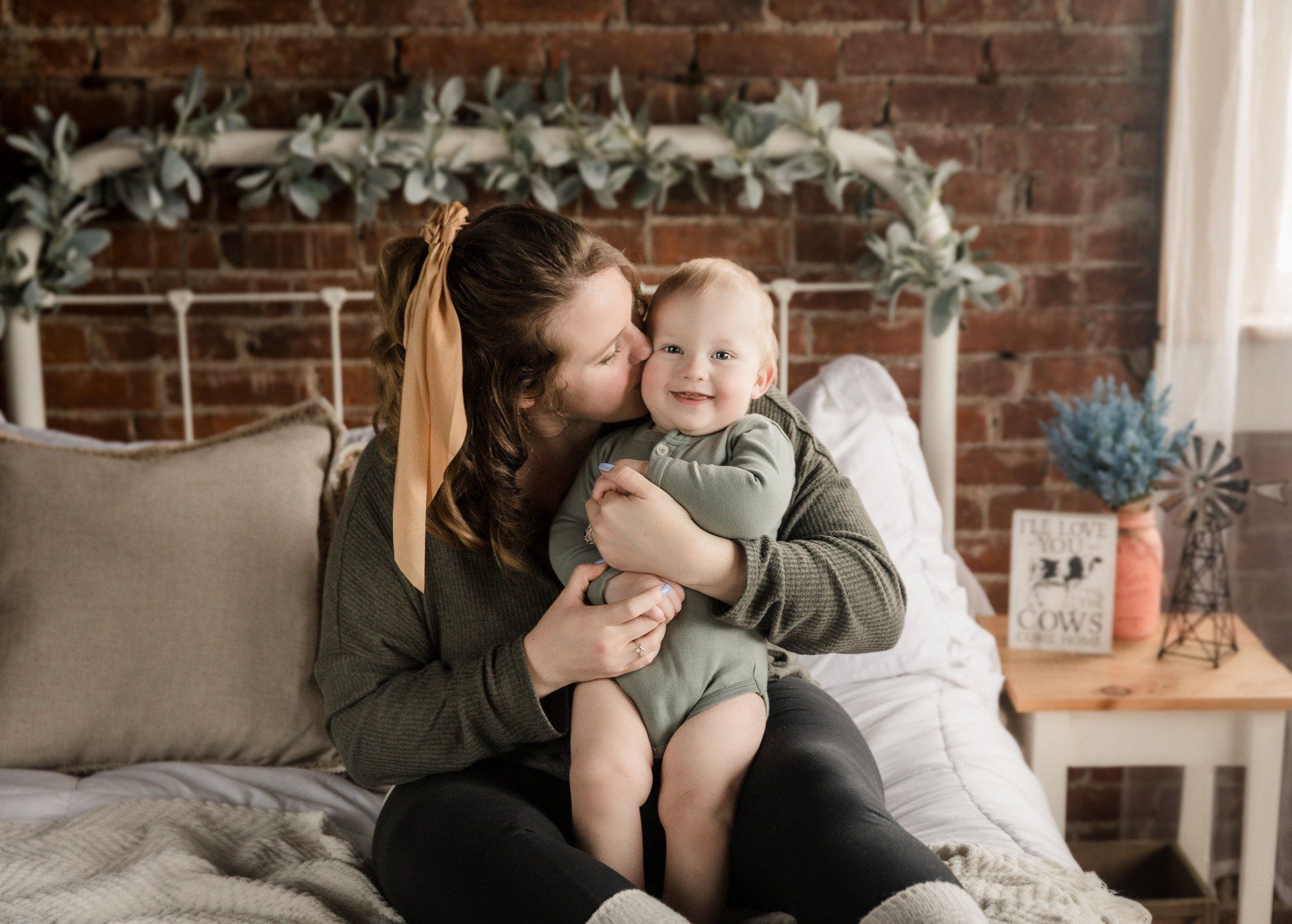 A woman is sitting on a bed holding a baby and kissing it on the cheek. Family photos