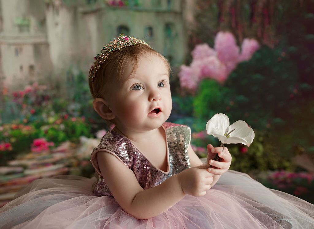 A baby girl in a pink dress and tiara is holding a flower.