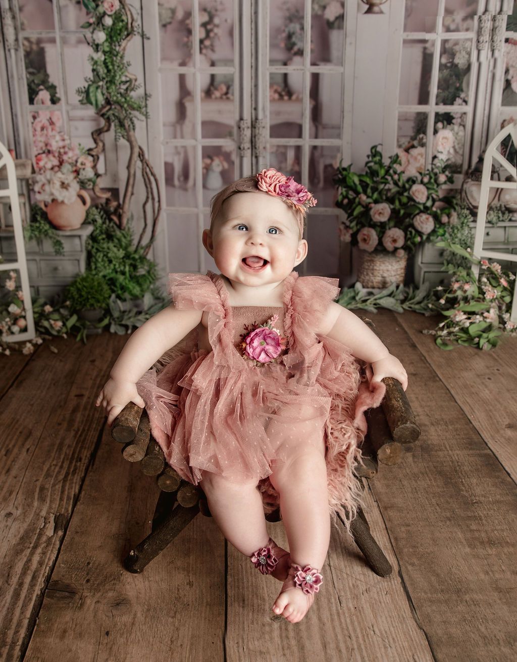 A baby girl in a pink dress is sitting in a wooden chair.