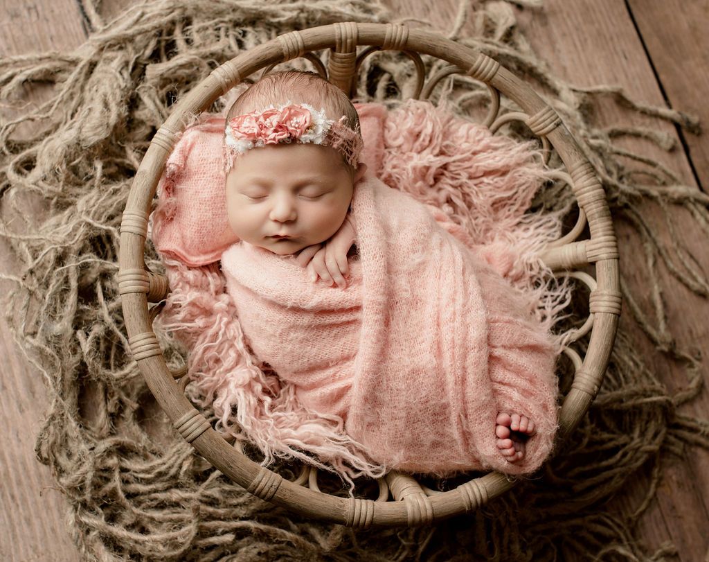 A newborn baby wrapped in a pink blanket is sleeping in a wicker basket.