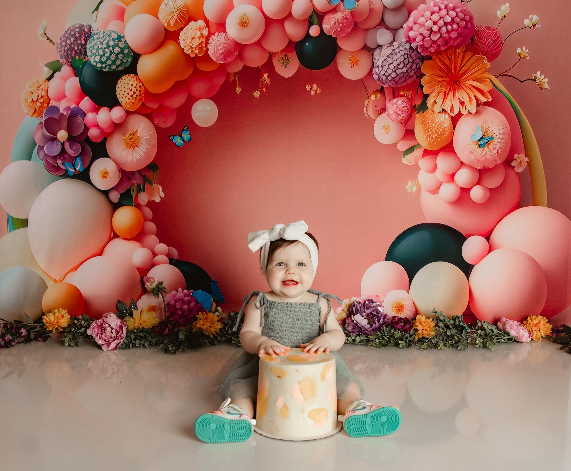 A baby is sitting in front of a balloon arch holding a cake.