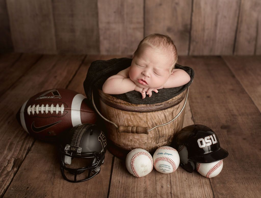 A newborn baby is sleeping in a bucket surrounded by footballs , baseballs , and a football helmet.