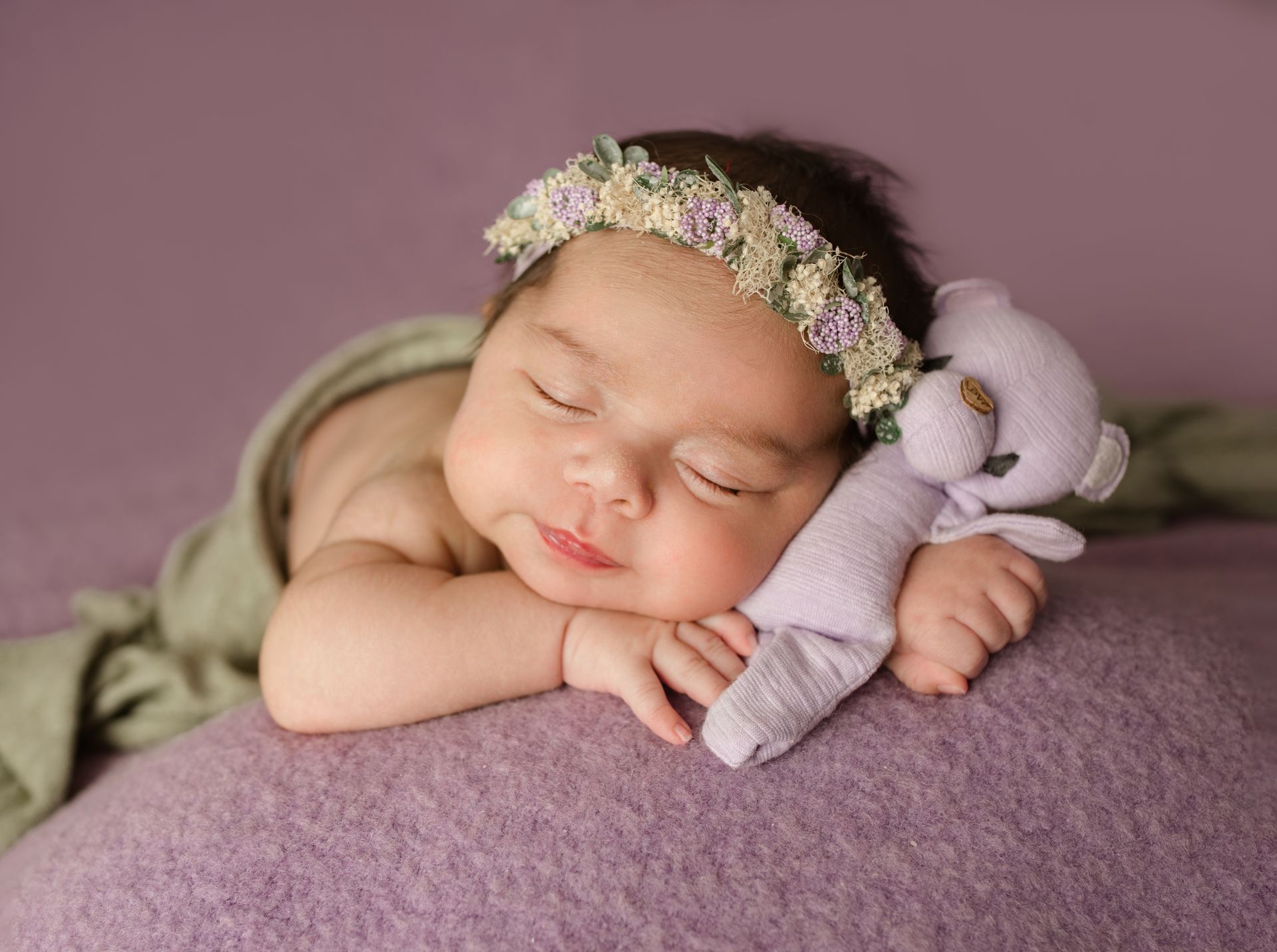 A newborn baby is sleeping on a purple blanket with a teddy bear.
