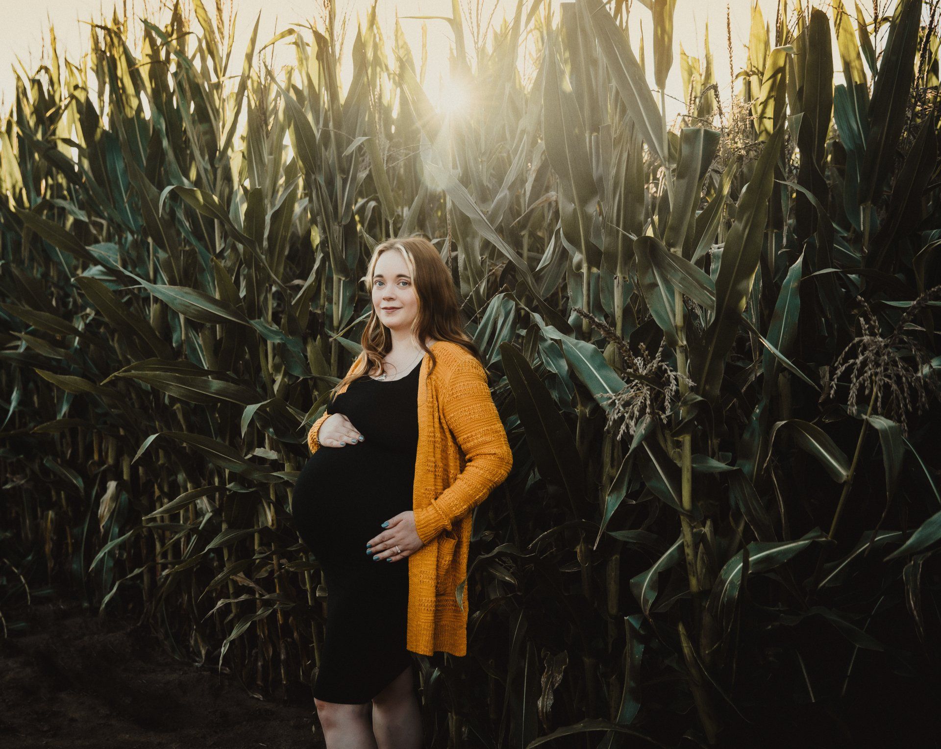 A pregnant woman is standing in a corn field holding her belly. Maternity Photographer near me