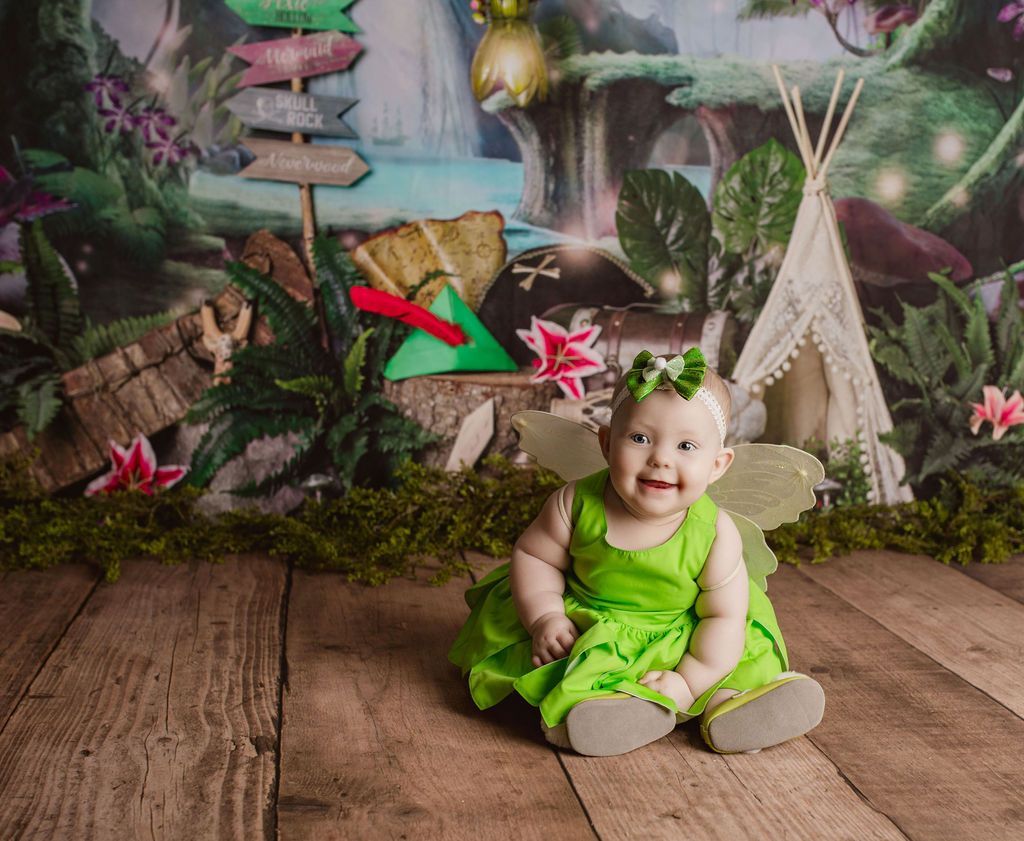 A baby in a green dress is sitting on a wooden floor.