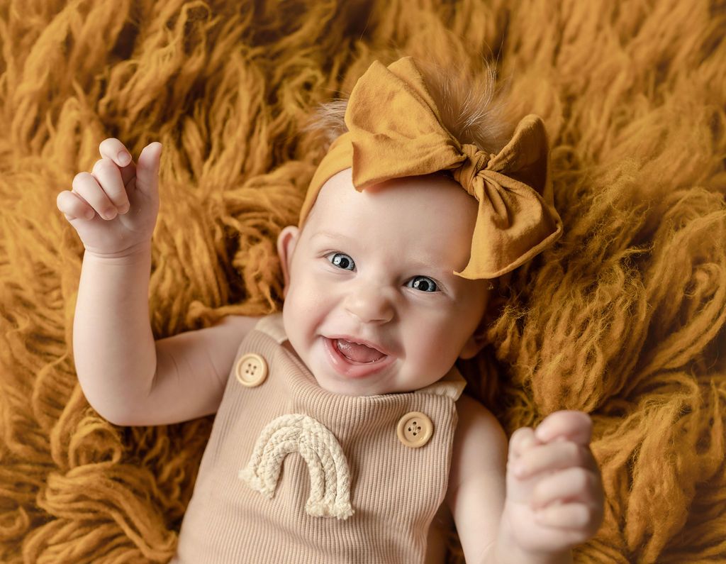A baby girl wearing a headband and overalls is laying on a furry blanket.