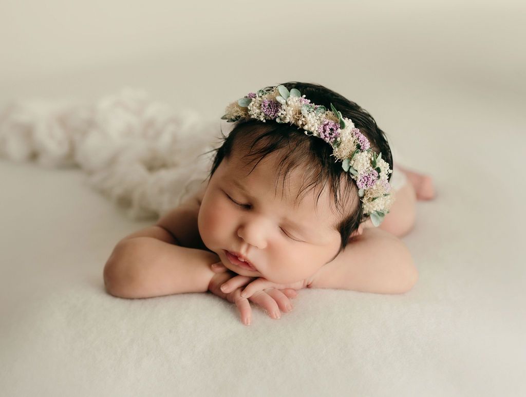 A newborn baby girl wearing a flower crown is sleeping on a bed.