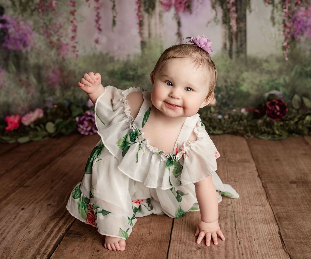 A baby girl in a white dress is crawling on a wooden floor.
