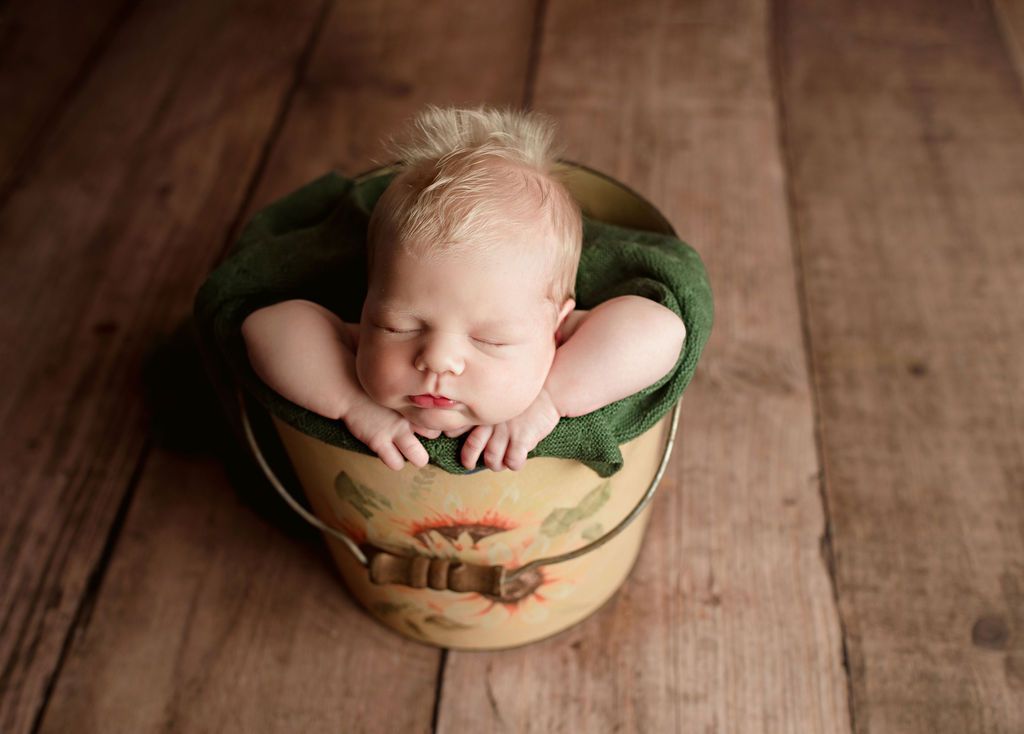 A newborn baby is sleeping in a bucket on a wooden floor.