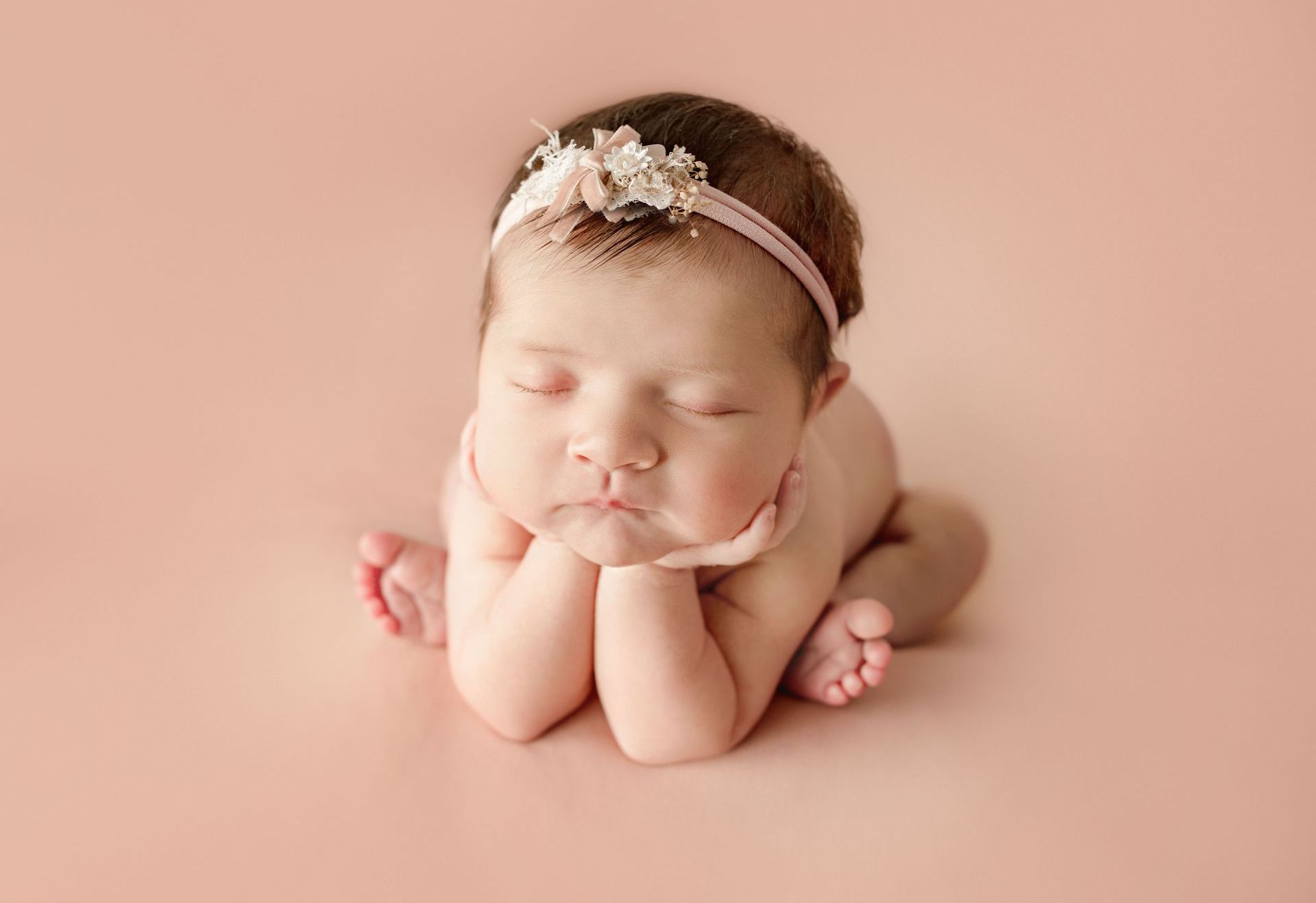 A newborn baby girl wearing a pink headband is sleeping on a pink background.