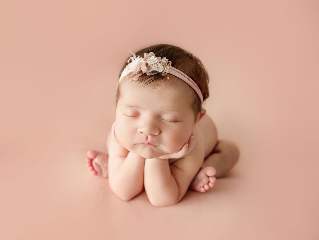 A newborn baby girl wearing a pink headband is sleeping on a pink background. Babys head in hands pose.