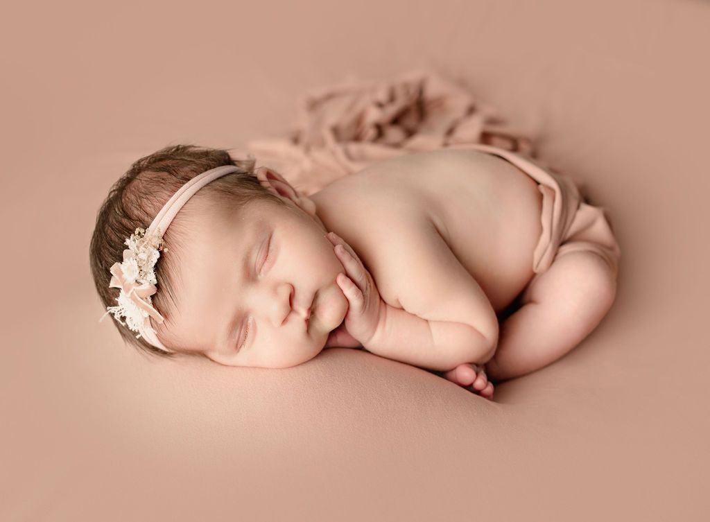 A newborn baby girl wearing a headband is sleeping on a pink blanket.