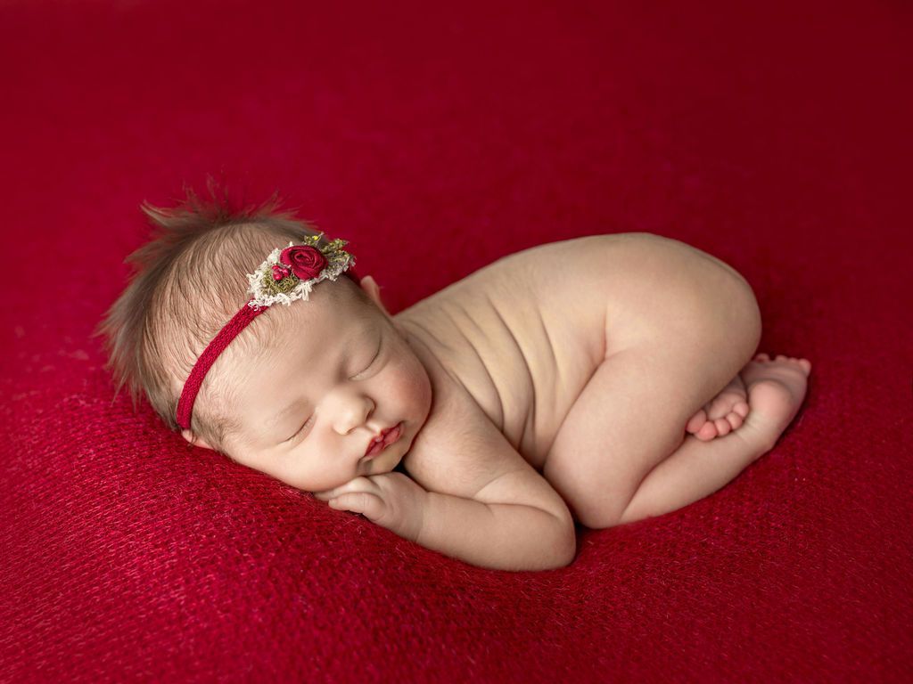 A newborn baby girl wearing a red headband is sleeping on a red blanket.