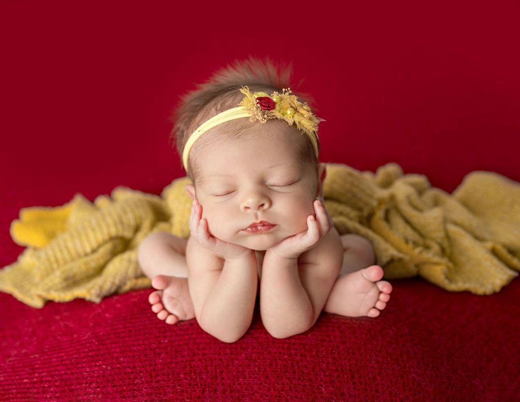 A newborn baby girl wearing a yellow headband is sleeping on a red blanket. Newborn head in hands pose
