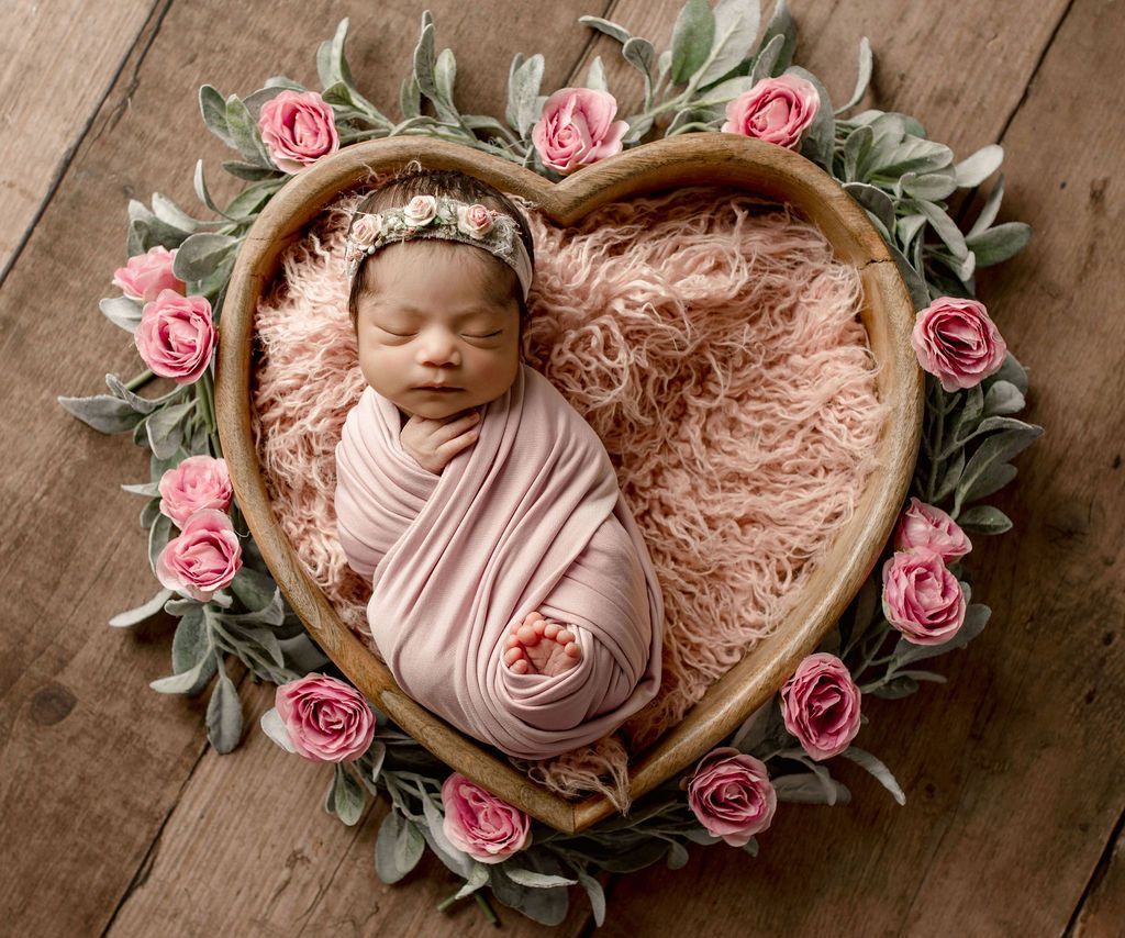 A newborn baby is sleeping in a heart shaped basket surrounded by pink flowers.
newborn baby studio