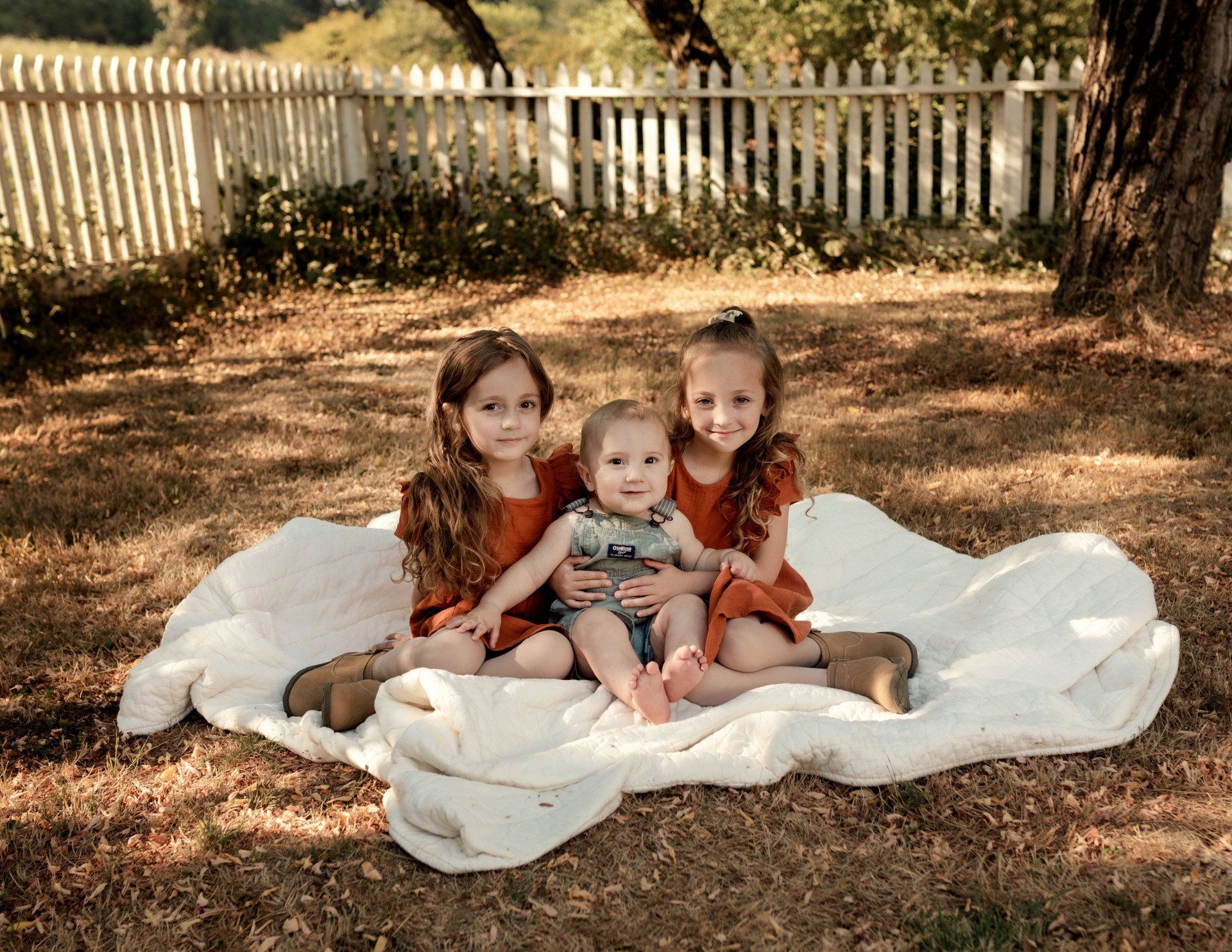 Three little girls are sitting on a blanket with a baby.