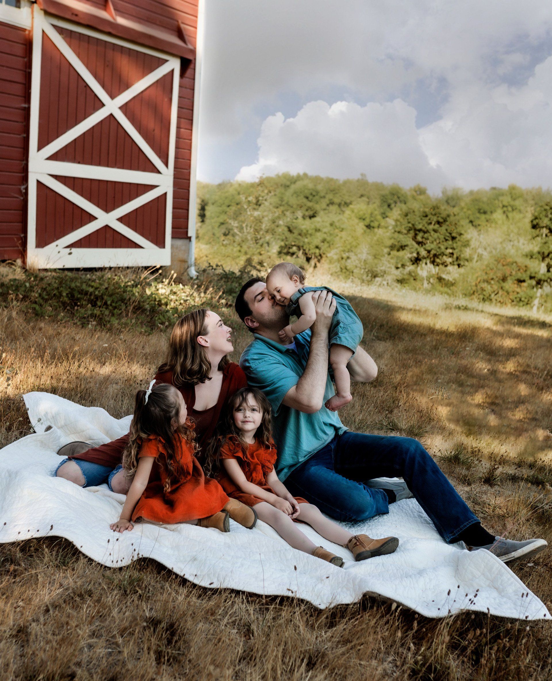 A family is sitting on a blanket in front of a barn.