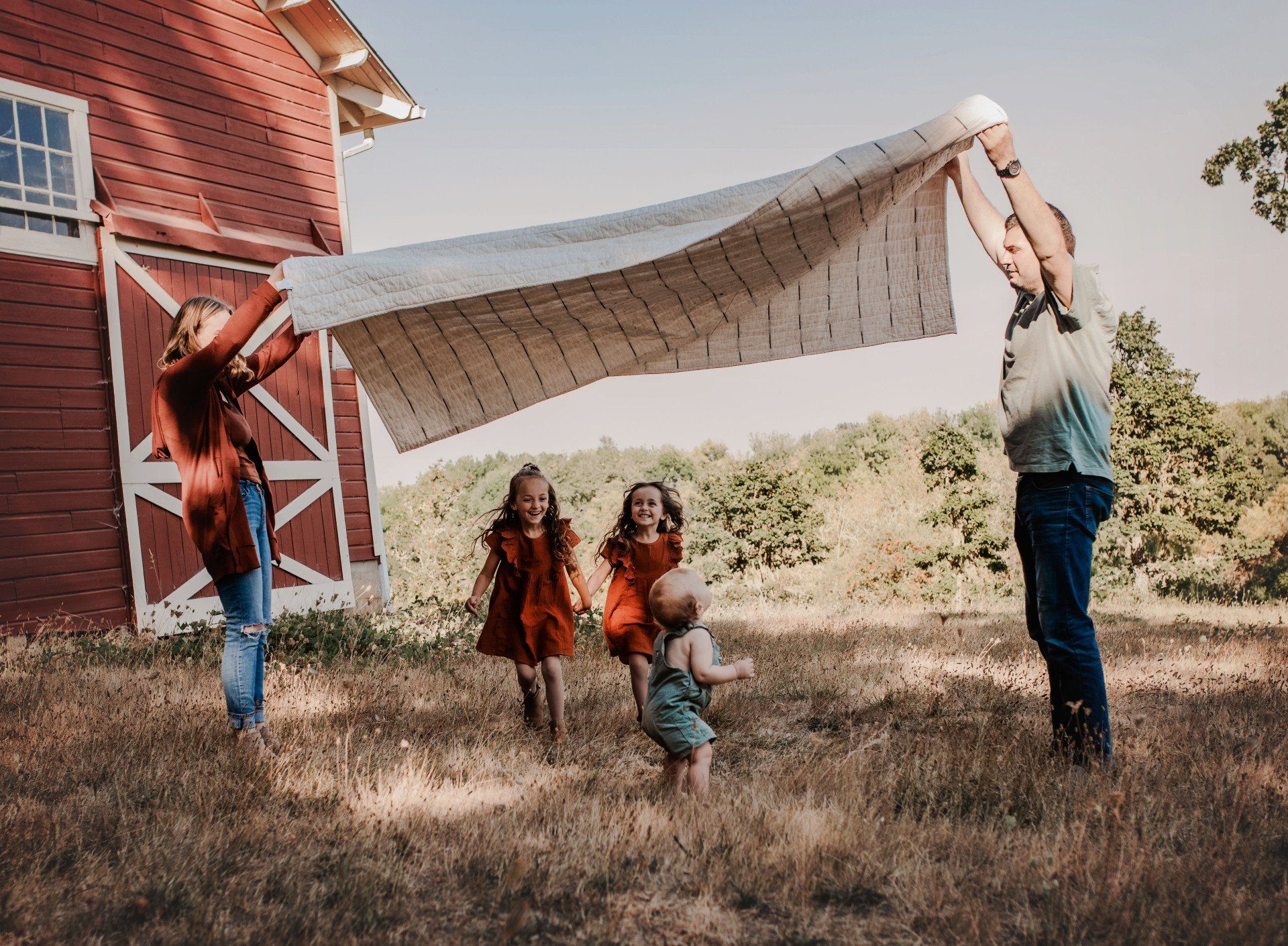 A family is playing with a blanket in front of a barn.