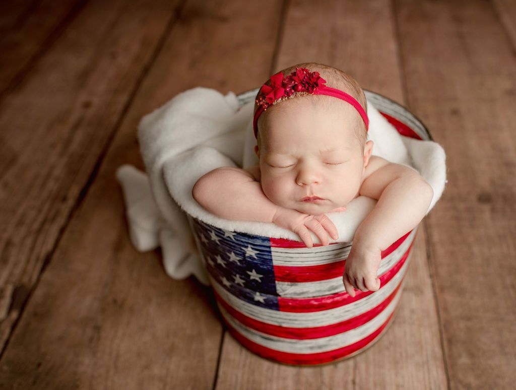 A newborn baby is sleeping in an american flag bucket.