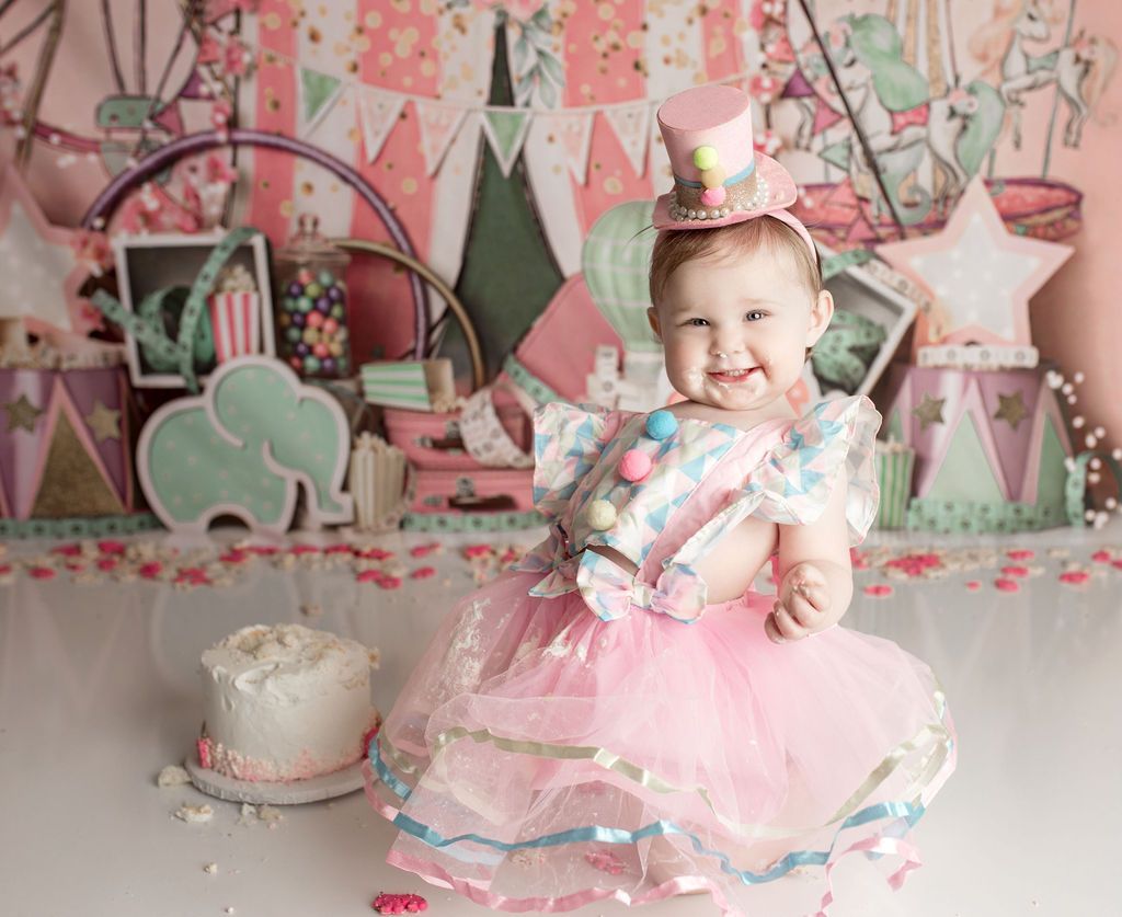 A baby girl in a pink dress and top hat is standing in front of a cake.