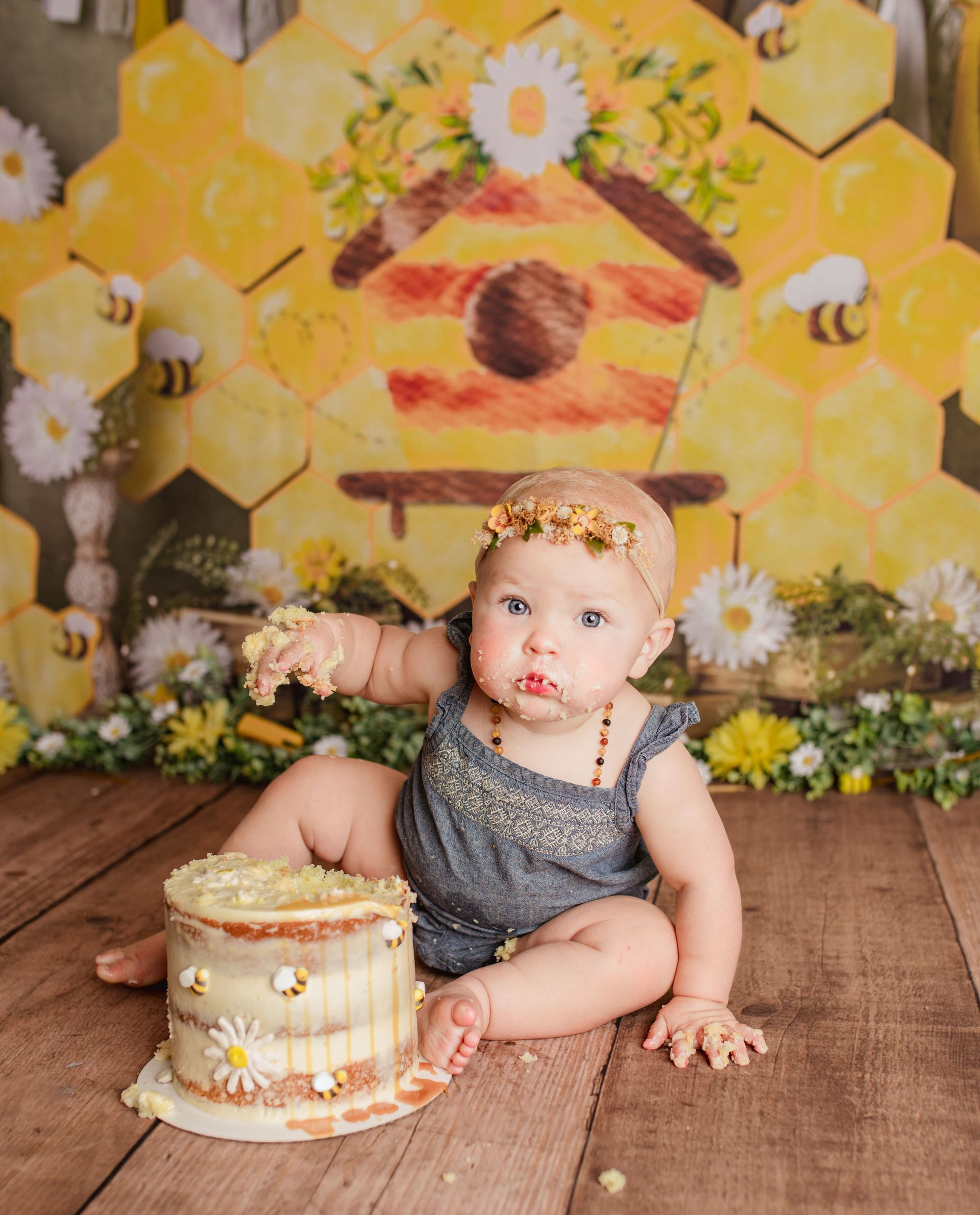 A baby is sitting on a wooden floor with a cake in front of a beehive.