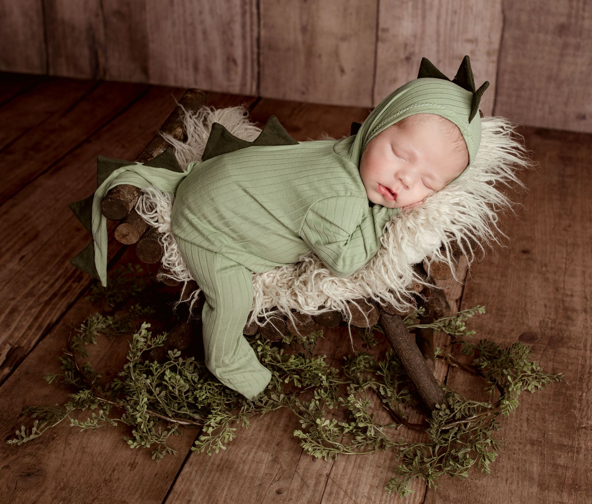A newborn baby wrapped in a green dragon costume is sleeping on a wooden bed.