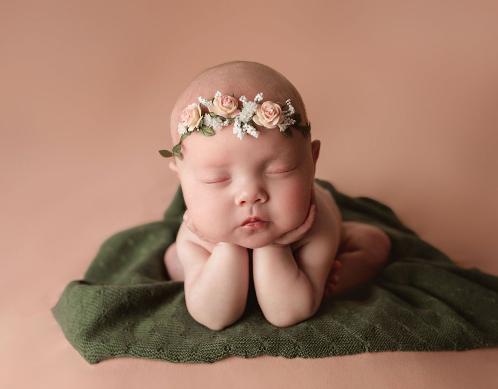 A newborn baby wearing a flower crown is sleeping on a green blanket.