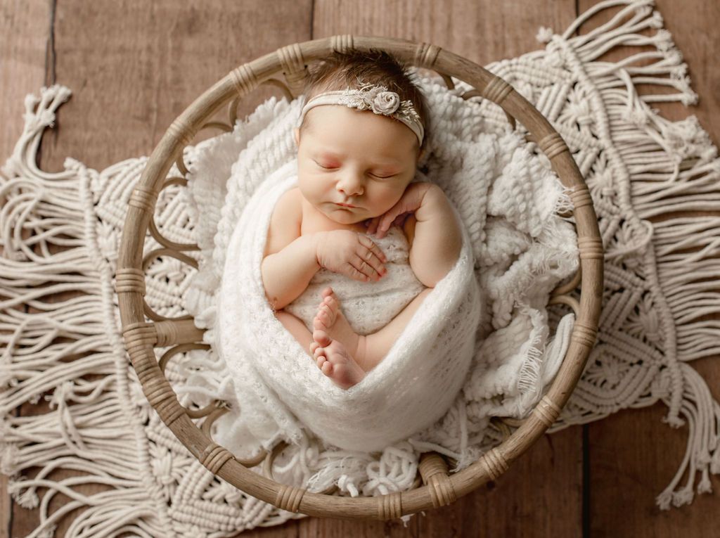 A newborn baby is wrapped in a white blanket and sleeping in a wicker basket. baby props