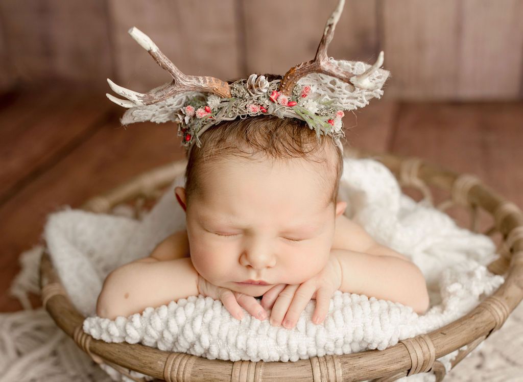 A newborn baby wearing a crown of antlers is sleeping in a basket.