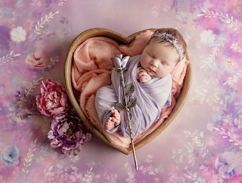 A newborn baby is sleeping in a heart shaped bowl with flowers.