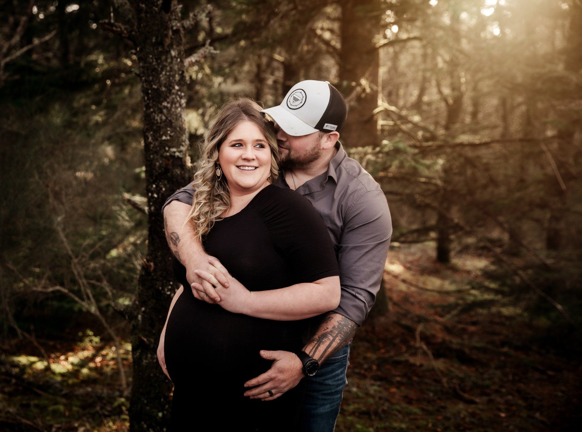 A man is kissing a pregnant woman on the cheek in the woods. Pregnancy photos