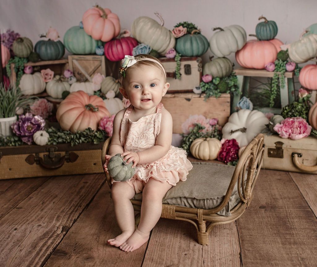 A baby girl is sitting on a wicker chair with pumpkins in the background.