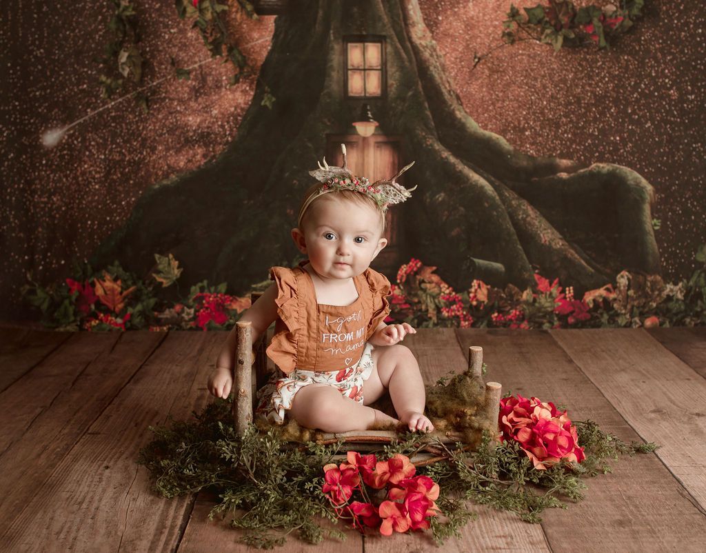 A baby girl is sitting on a wooden chair in front of a tree.