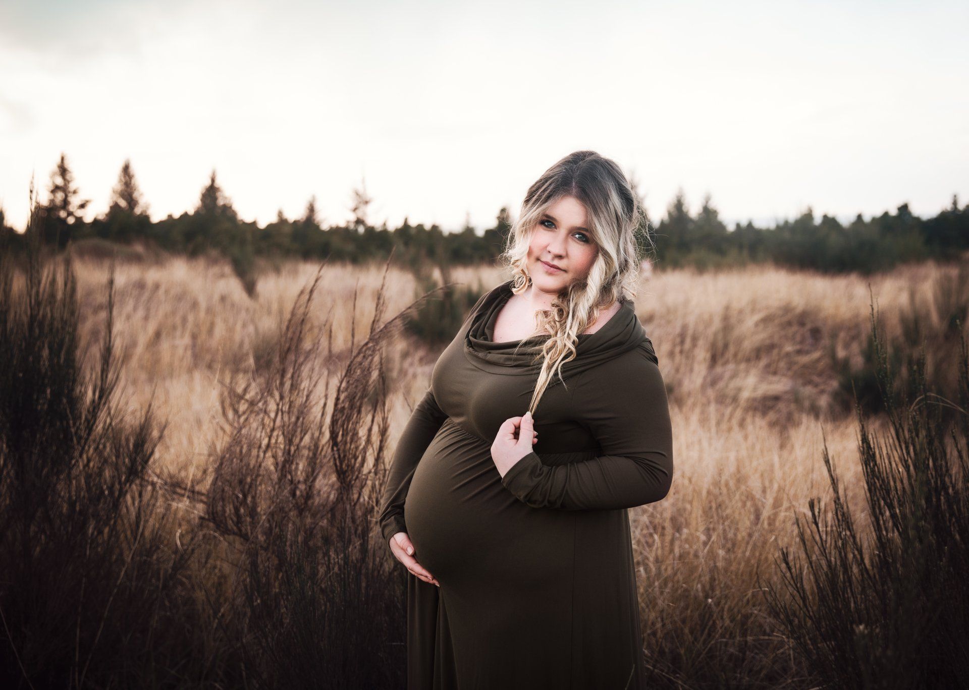 A pregnant woman in a green dress is standing in a field holding her belly.