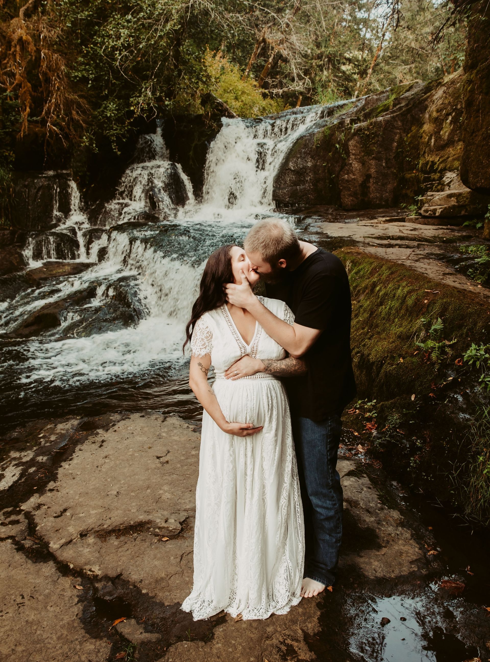 A man and a pregnant woman are kissing in front of a waterfall.