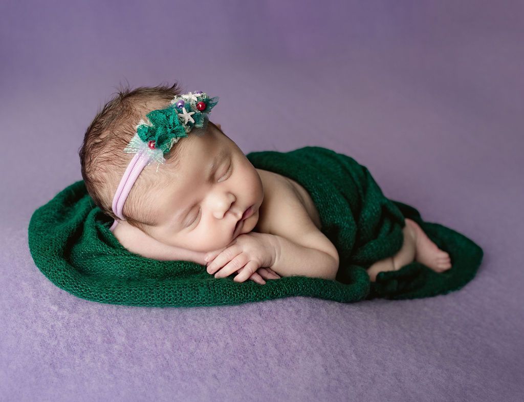 A newborn baby is wrapped in a green blanket and wearing a headband. newborn girl props