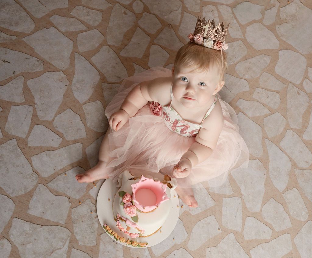 A baby girl wearing a crown is sitting next to a cake.