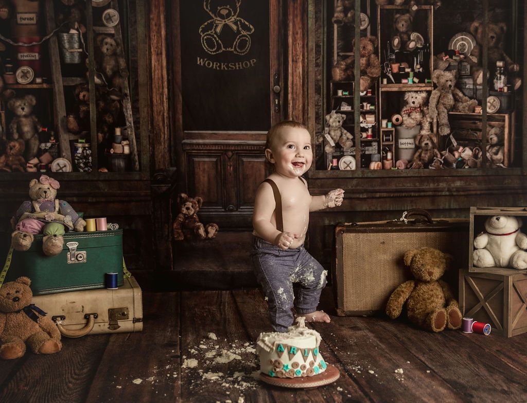 A baby is standing on a cake in front of a wall of teddy bears.