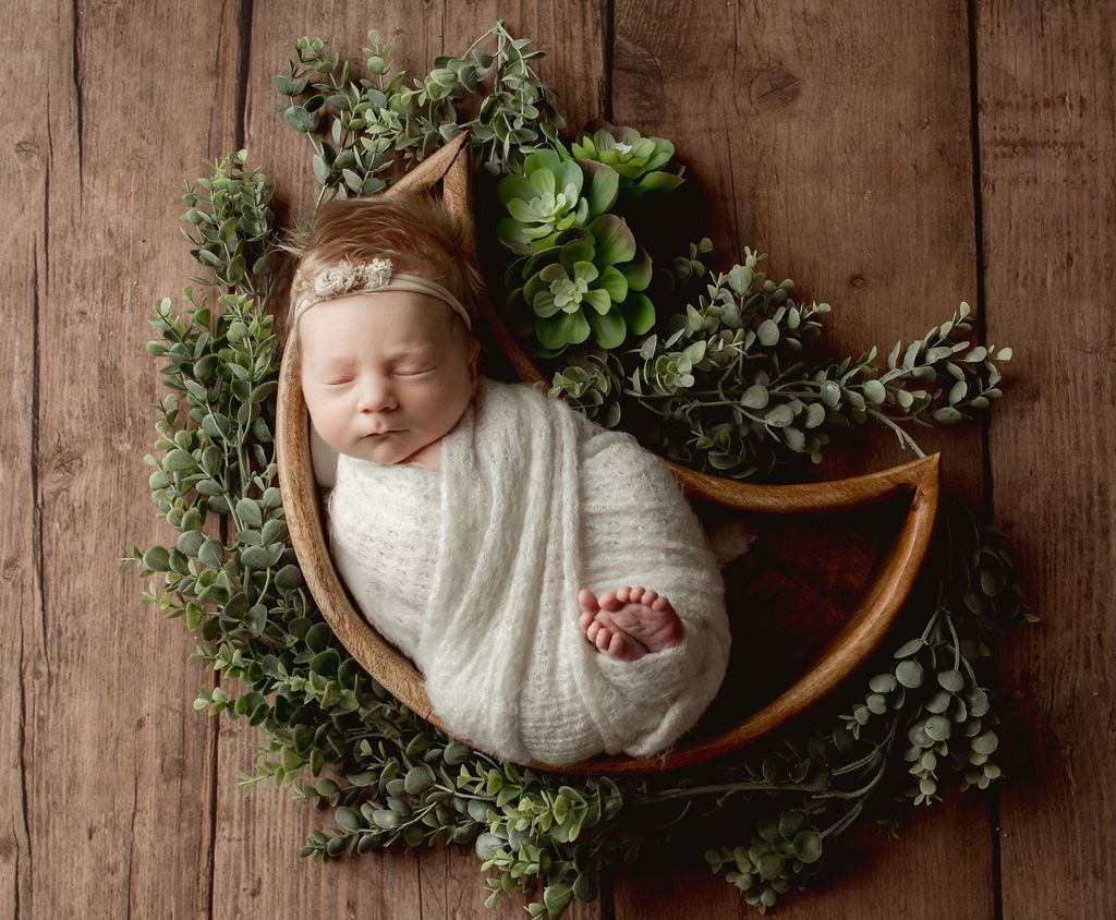 A newborn baby wrapped in a white blanket is sleeping in a wooden basket surrounded by greenery.