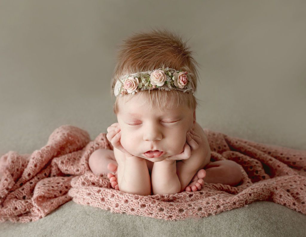 A newborn baby girl wearing a flower headband is sleeping on a pink blanket.