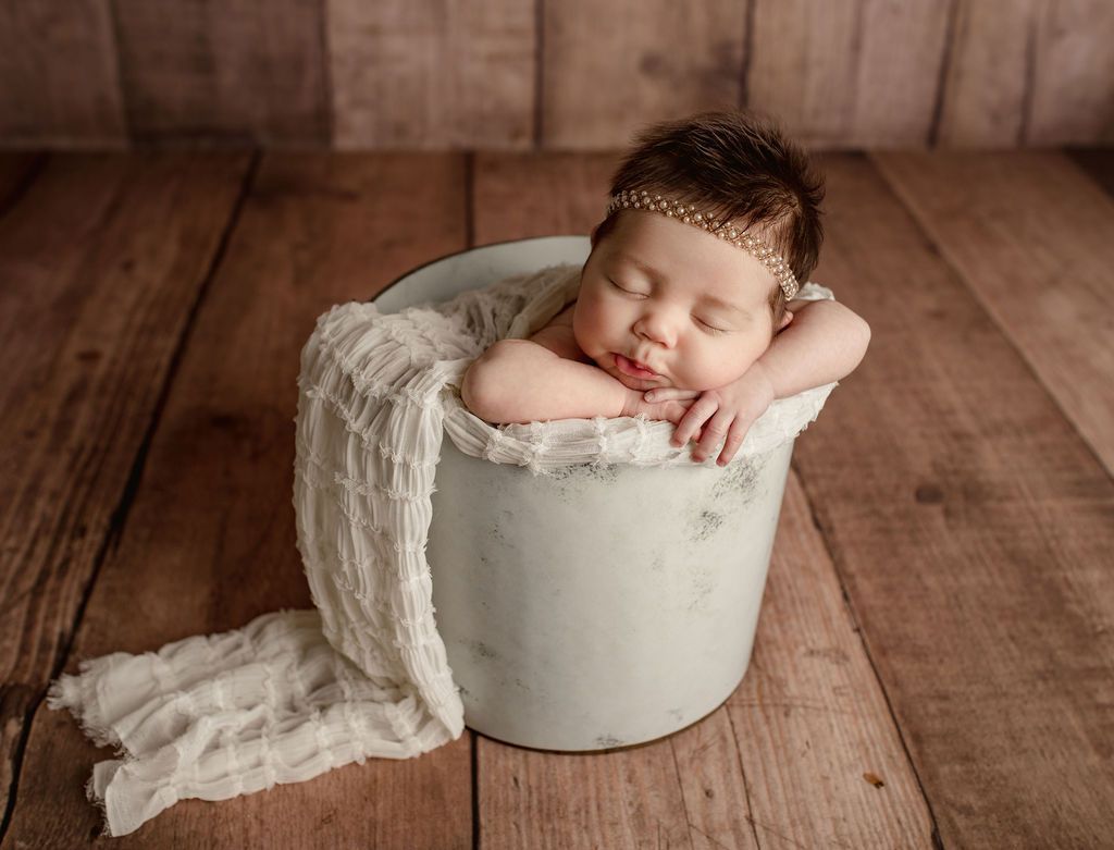 A newborn baby is sleeping in a white bucket on a wooden floor.