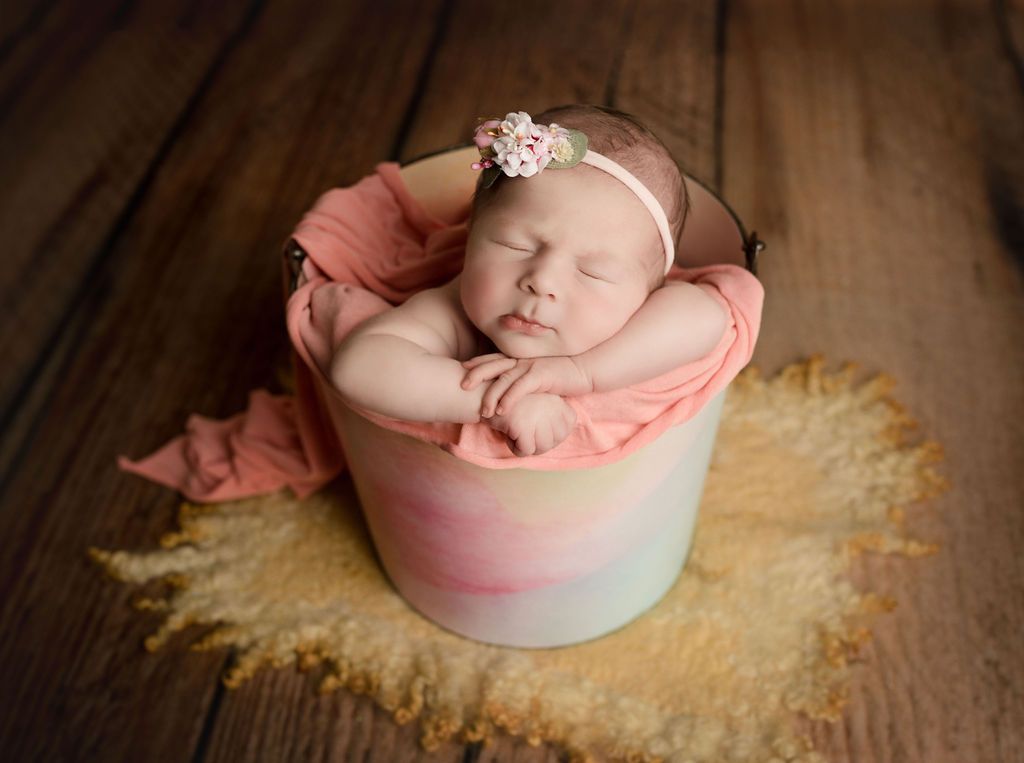 A newborn baby is sleeping in a bucket on a wooden floor.