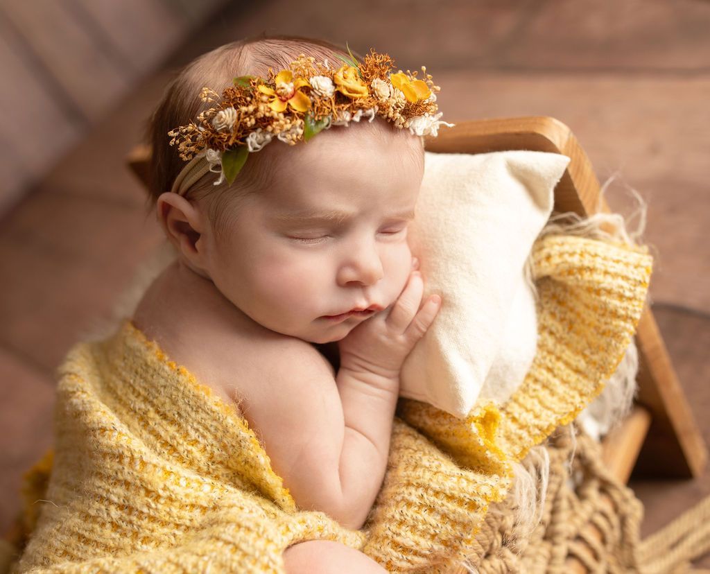 A newborn baby wearing a flower crown is sleeping in a basket.