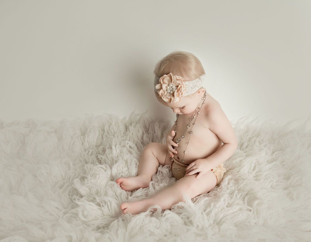 A baby girl wearing a headband and necklace is sitting on a white blanket.